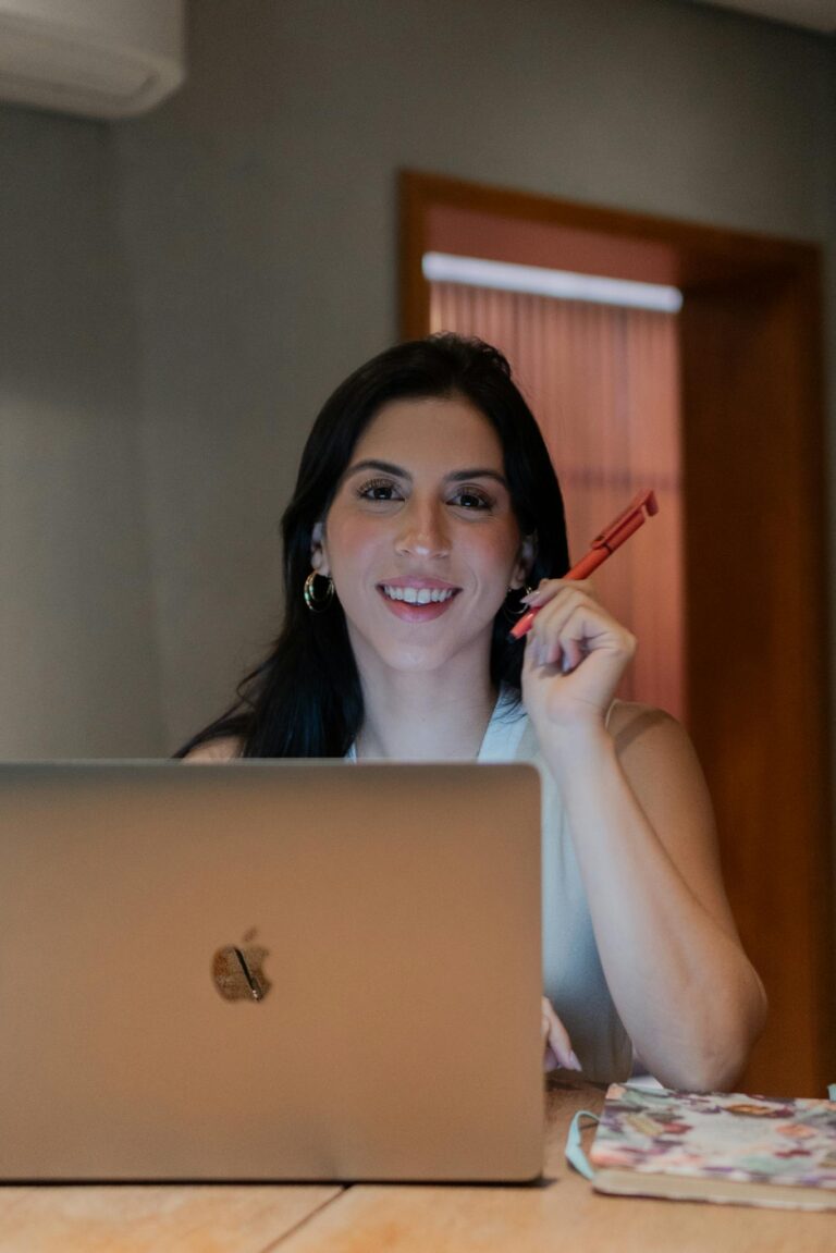 Smiling woman holding pen while working on laptop at desk.