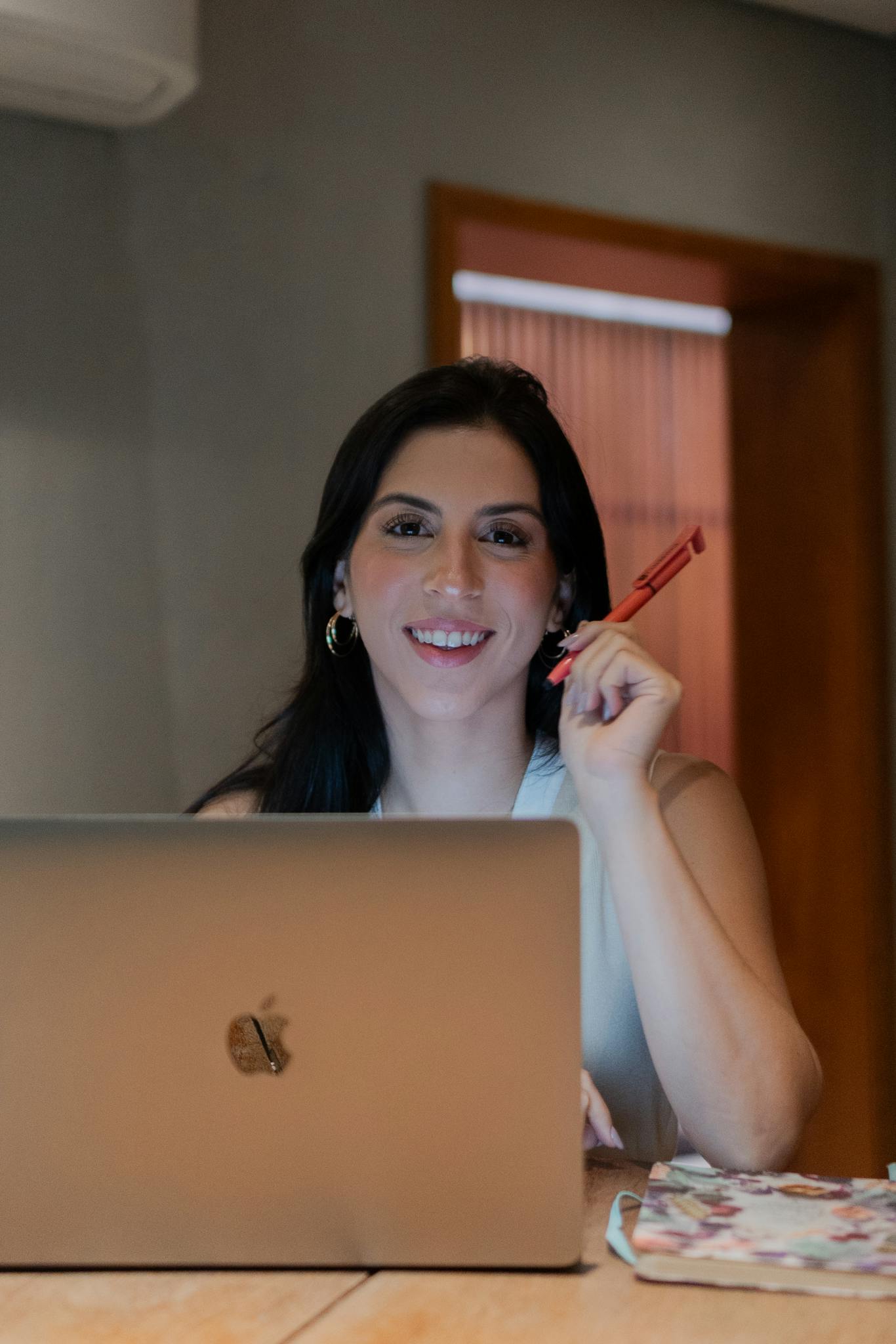 Smiling woman holding pen while working on laptop at desk.