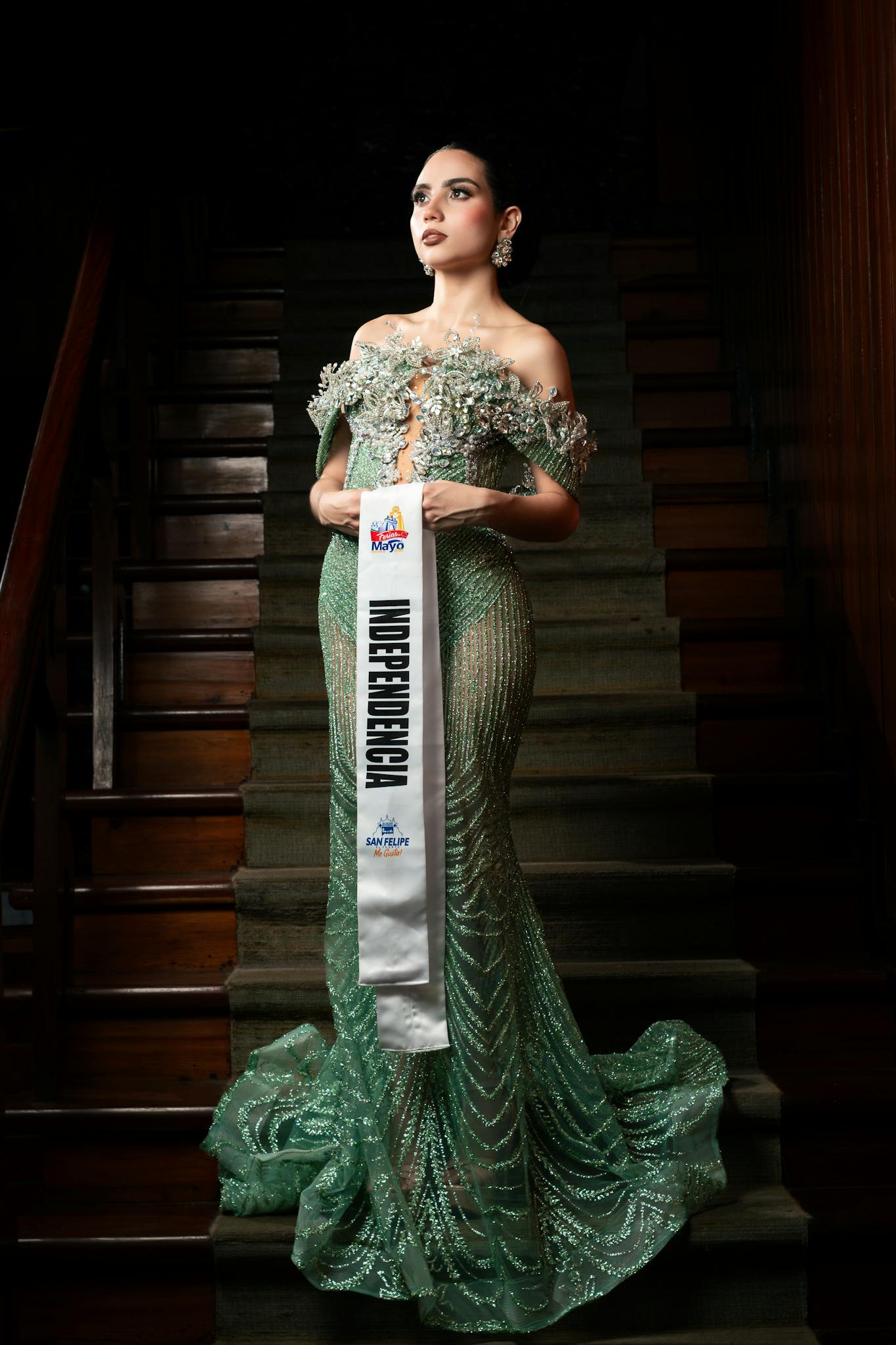 Woman in glamorous gown posing on staircase with contest sash indoors.