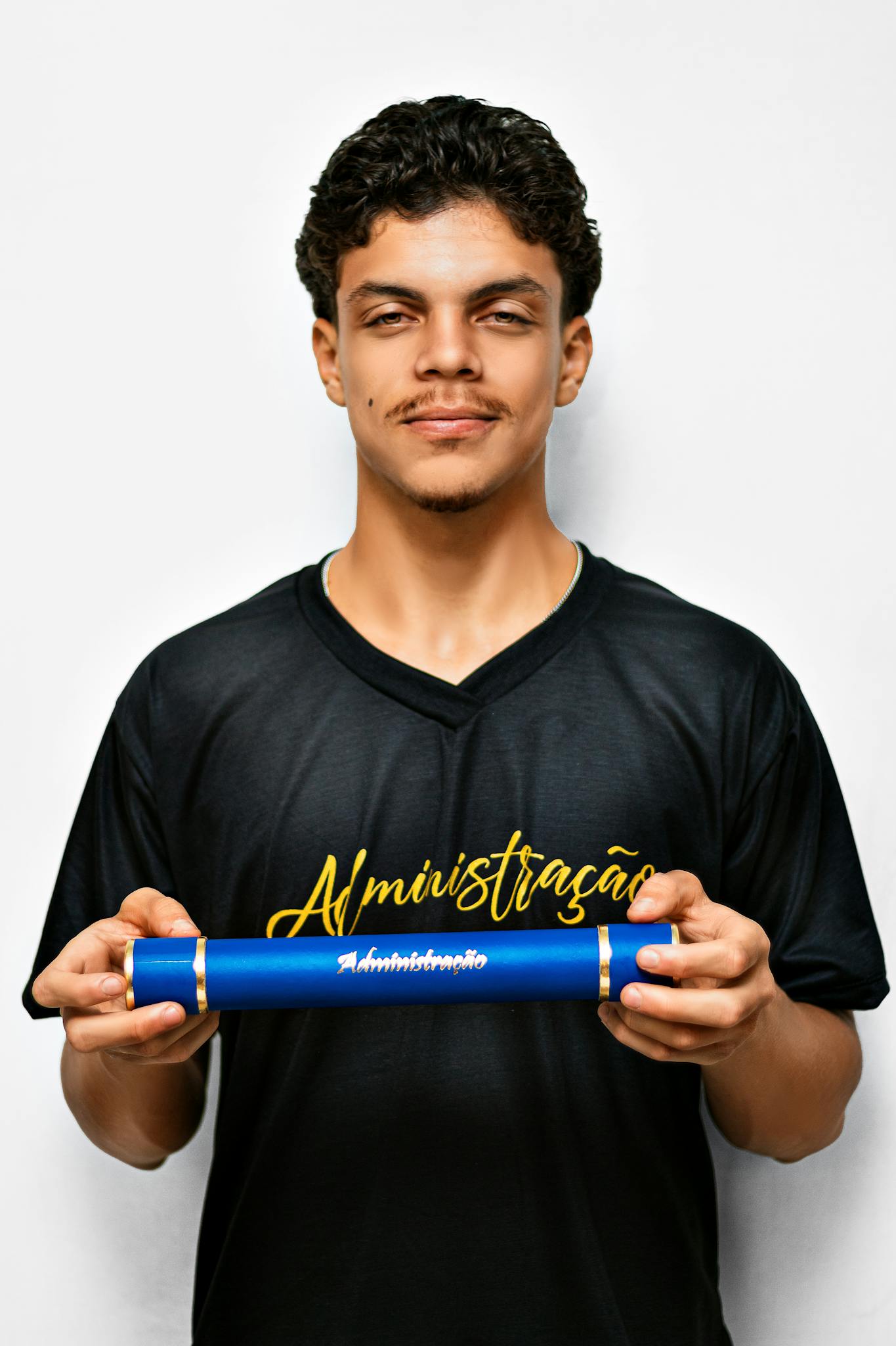 Young adult wearing black shirt holding a blue diploma tube against a white background.